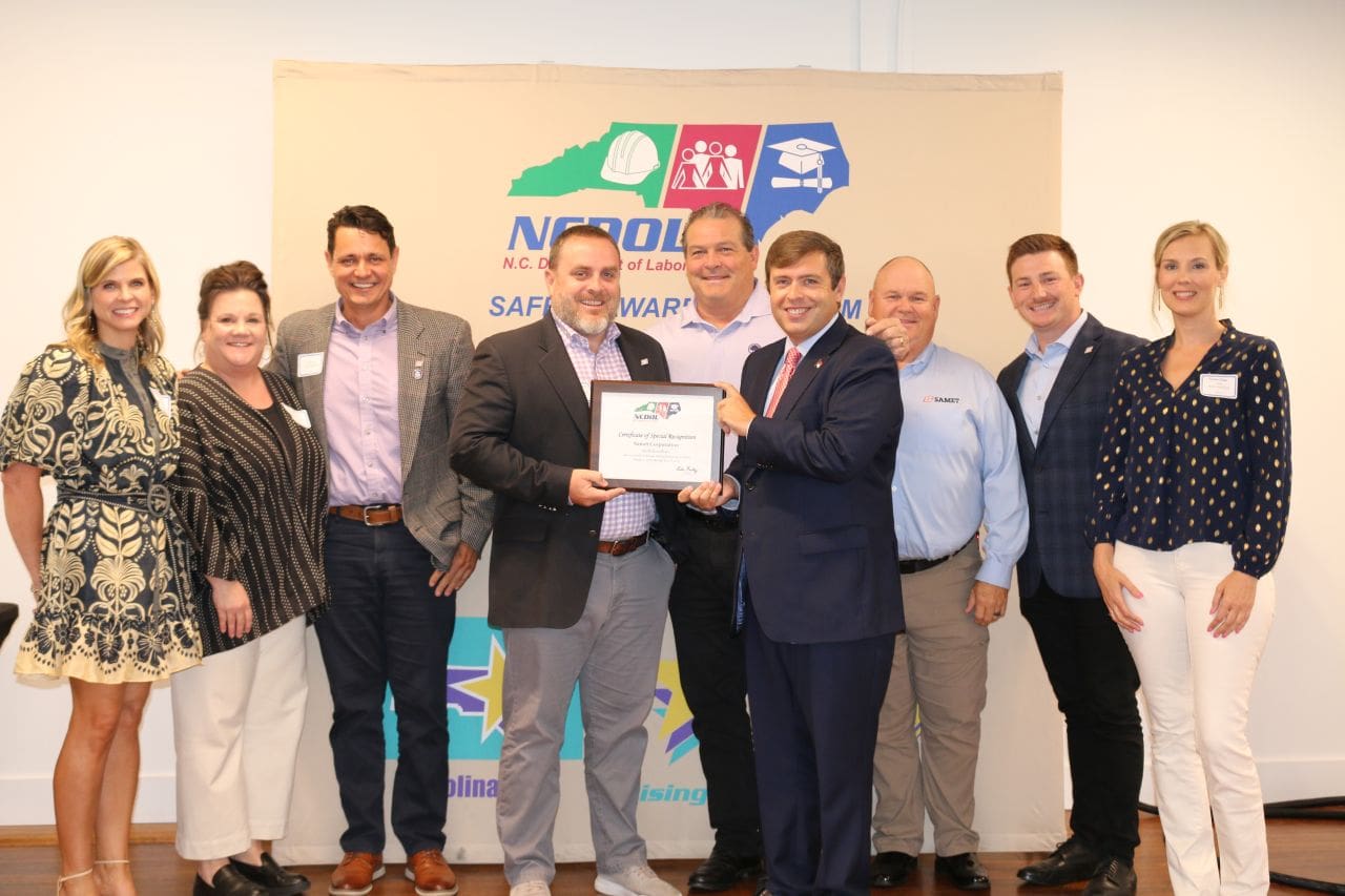 Two representatives from Samet Corporation stand with a North Carolina Department of Labor official while receiving a safety award, smiling in front of a backdrop featuring NCDOL branding.