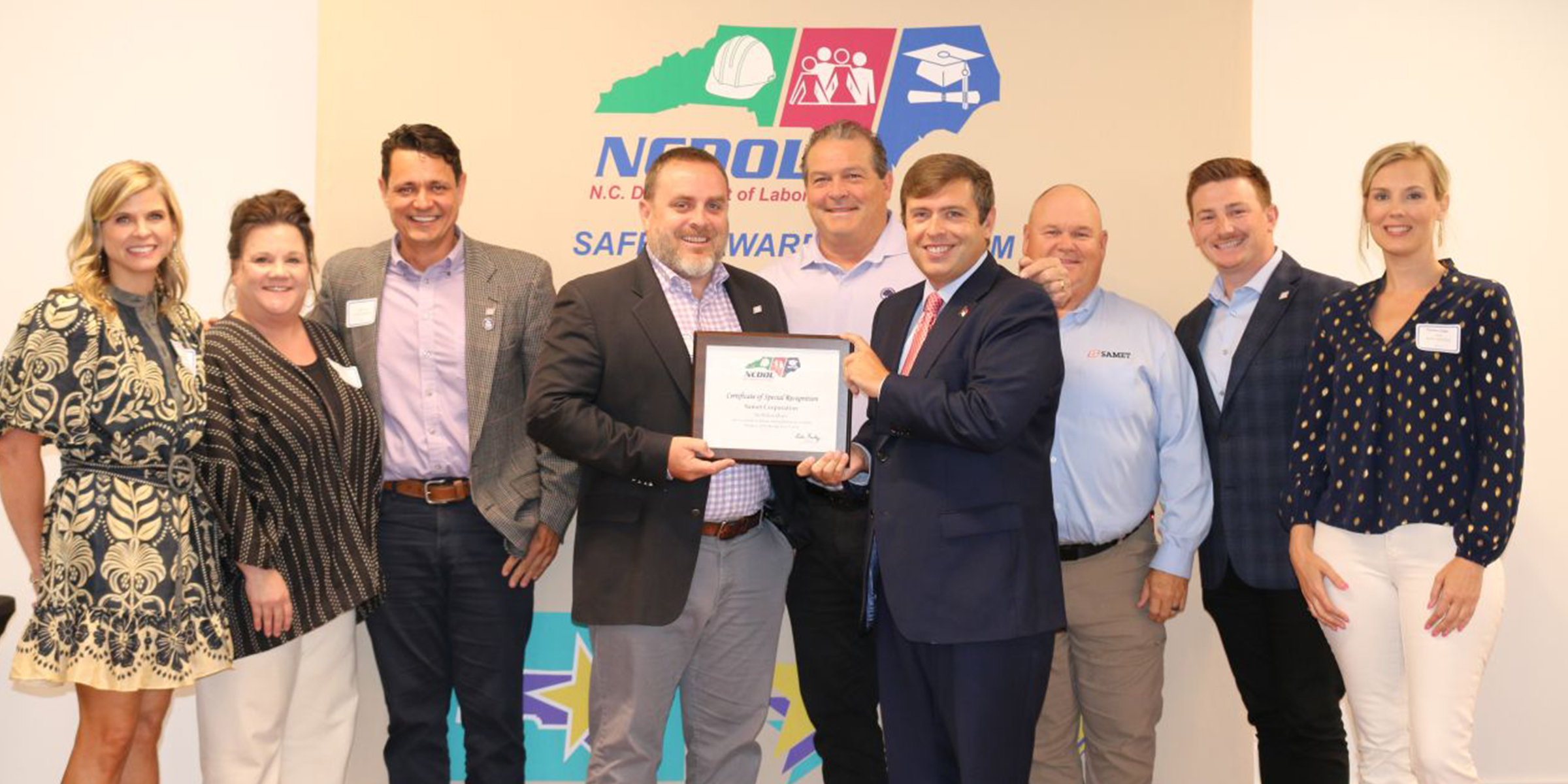 Two representatives from Samet Corporation stand with a North Carolina Department of Labor official while receiving a safety award, smiling in front of a backdrop featuring NCDOL branding.